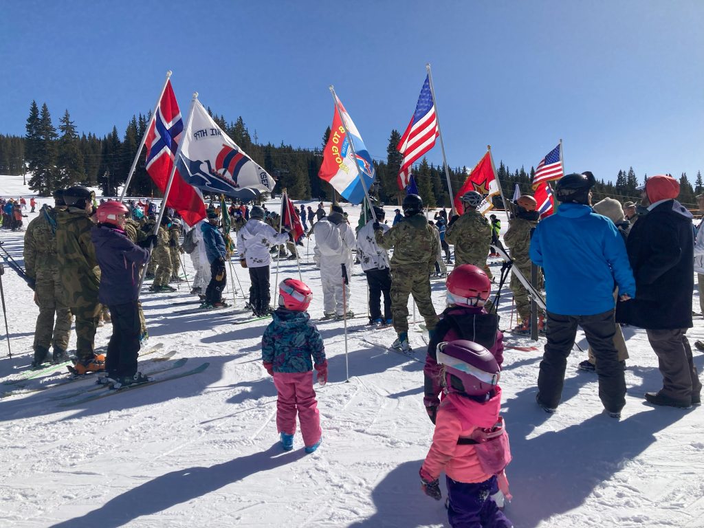 Skiers gather on a snow-covered slope, holding various national flags, including American and Norwegian. The scene is lively, with children in colorful winter gear.
