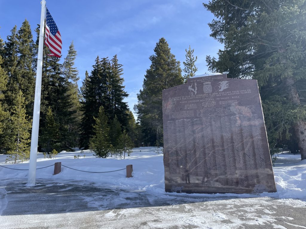 A memorial plaque stands under a clear blue sky, surrounded by snow and evergreen trees. An American flag is flying on a pole beside it. The scene is calm and reflective.