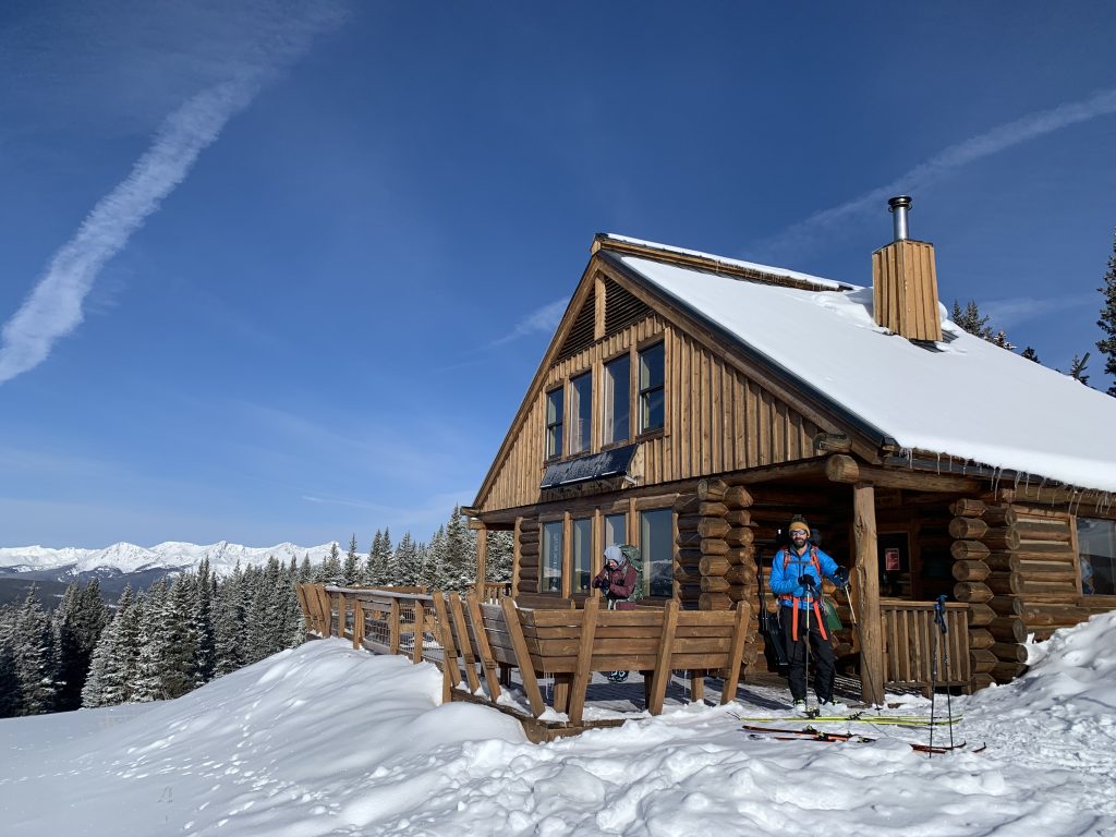 Log cabin in snowy landscape with a clear blue sky. Two people in winter gear stand outside, overlooking snow-covered trees and distant mountains.