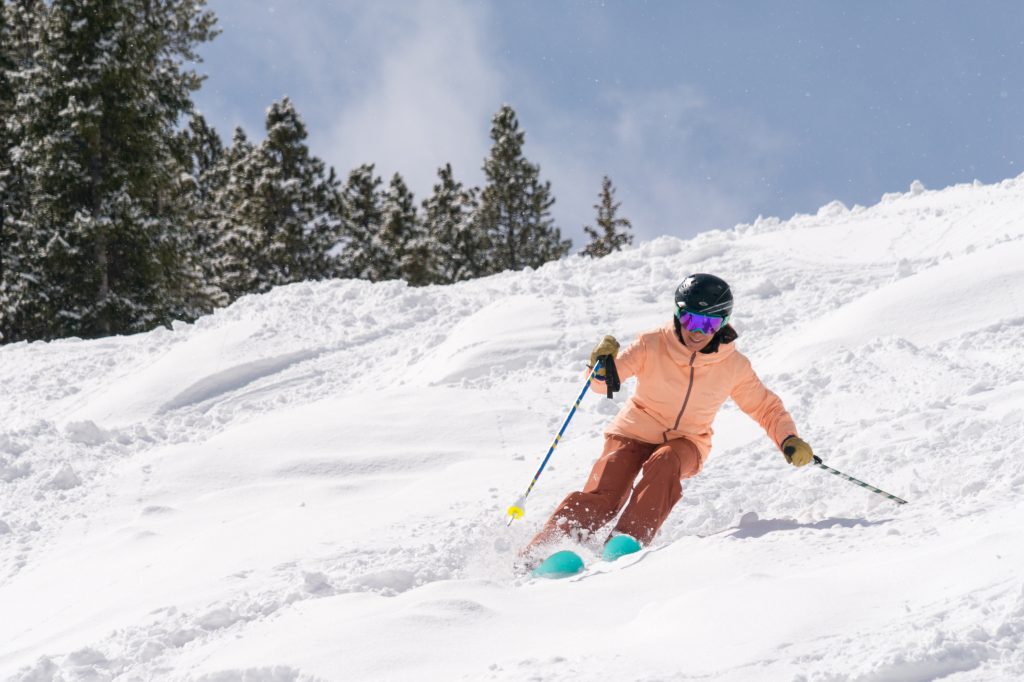 A skier in an orange jacket skillfully navigates a snowy slope, surrounded by tall pine trees under a clear sky, evoking excitement and adventure.
