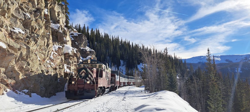 A vintage train travels along a snow-covered track in a mountainous forest, with rocky cliffs on the left. The sky is bright blue with wispy clouds.