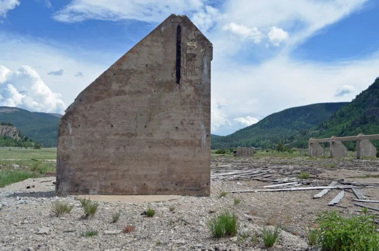 A solitary, weathered concrete wall, once part of a military base, stands amid scattered debris on a barren landscape, under a bright blue sky and fluffy clouds, evoking a sense of desolation.