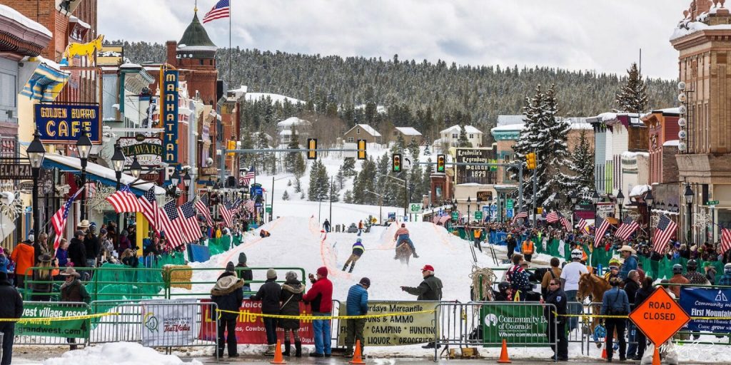 A vibrant outdoor winter festival with snowy street, skiers racing downhill. Lined with crowds, American flags, colorful storefronts; festive atmosphere.