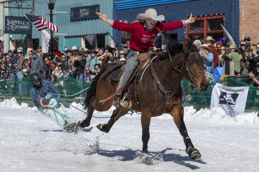 Rider on a galloping horse pulls a skier through snowy streets during a skijoring event. Enthusiastic crowd watches, waving flags in the background.