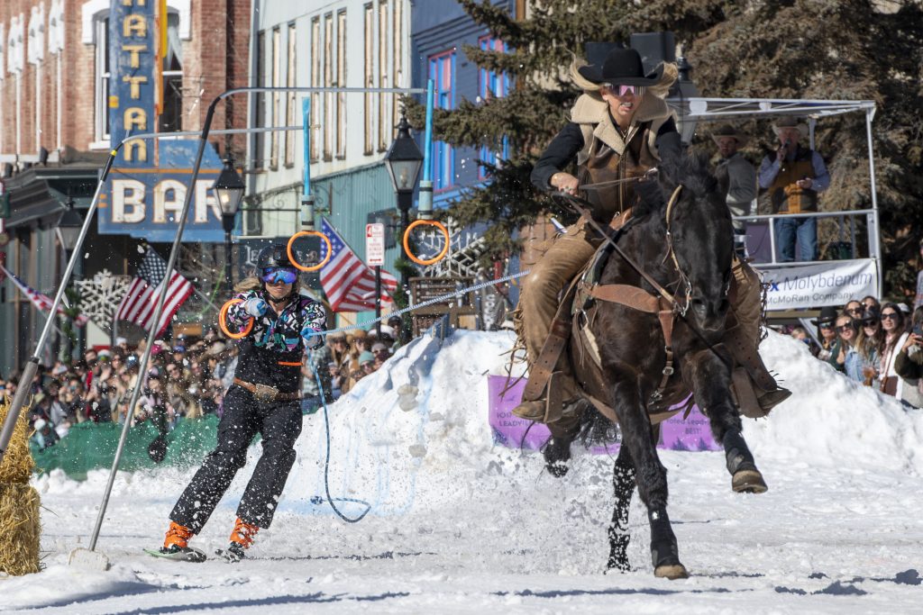 A skier being pulled by a cowboy on horseback in a snowy, lively street event. The skier grabs rings while spectators watch, conveying excitement and action.