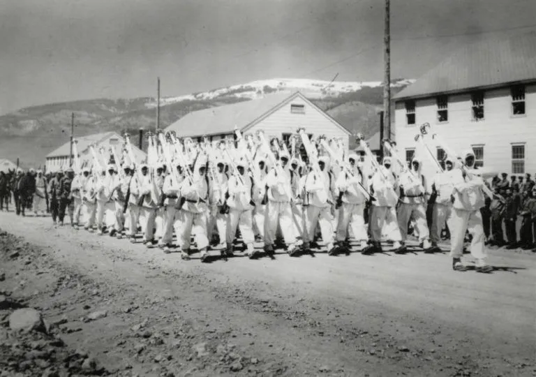 A 10th Mountain Division unit of soldiers marches in parade formation. All are wearing camouflage winter “whites” and carry skis on their right shoulders