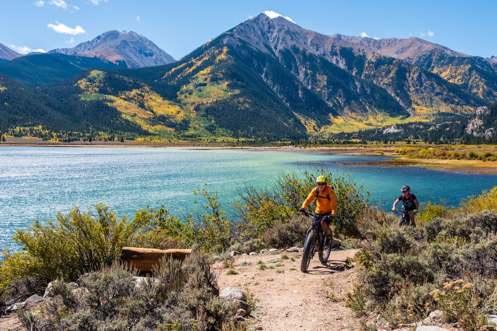 people biking on the side of a lake with mountains in the background.