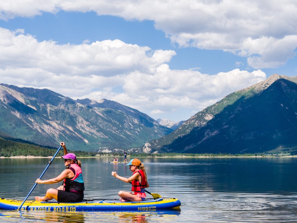 two people on paddle bored on a lake with mountains in the background.