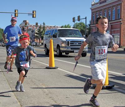photo of city street with two children and an adult running in a race, numbers pinned to their shirts. Yellow cones and a white van in the street behind them.