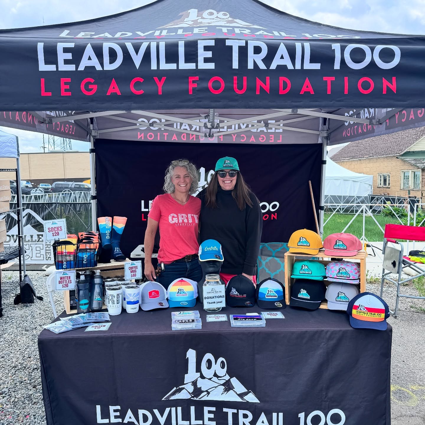 photo of a black expo booth tent for the Leadville Trail 100 Foundation. Two women stand behind a table with a black logoed tablecloth and hats, socks and water bottles for sale.