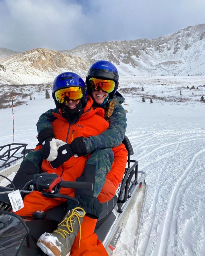 Two people in bright snow gear and helmets are joyfully riding a snowmobile in a snowy mountain landscape. Tracks are visible in the snow.
