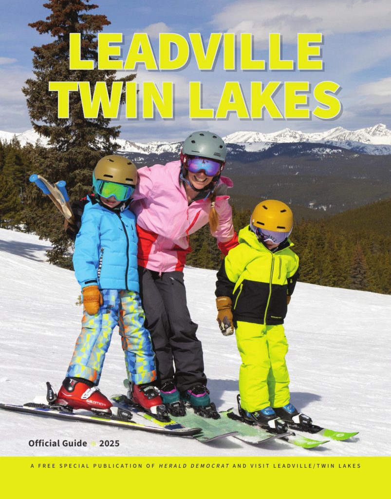 A family of three skiing on a snowy mountain. They're smiling, wearing bright outfits and helmets. Snowy peaks and pine trees are in the background.