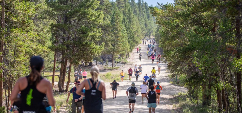 Trail runners are spread out on a dirt path surrounded by dense green forest, under clear skies. The scene conveys energy and determination.