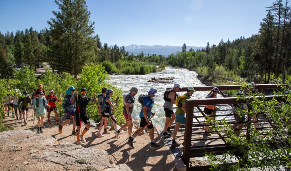 Group of trail runners crossing a wooden bridge over a rushing river in a forested area. The scene is vibrant, energetic, and sunny.