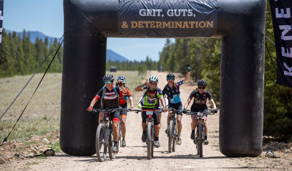 Five cyclists joyfully cross the finish line under an archway reading "Grit, Guts, & Determination" on a dirt trail surrounded by trees and mountains.