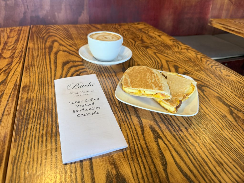 A menu, latte, and pressed sandwich sit on a rustic wooden table. The inviting cafe setting suggests a cozy, relaxed atmosphere.