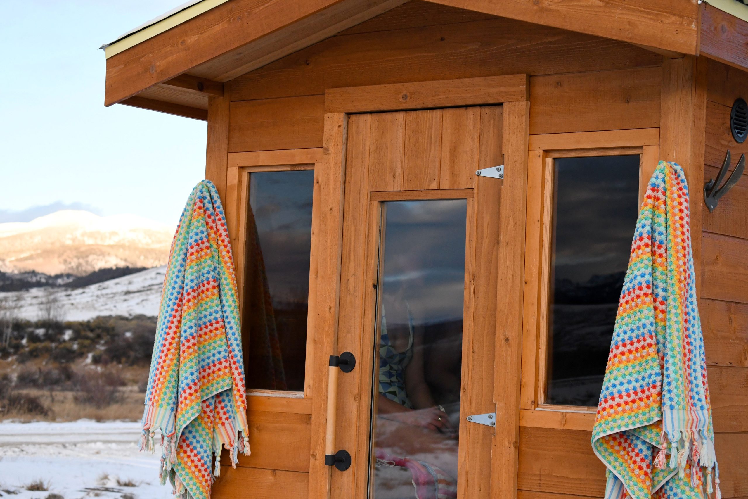 A small wooden sauna with colorful towels hanging on either side of the door stands against a snowy mountain backdrop, creating a cozy and serene winter scene.