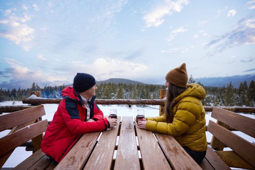 Two people in winter coats sit across a wooden picnic table, outdoor snowy landscape in the background. They smile, holding glasses, beneath a cloudy sky.