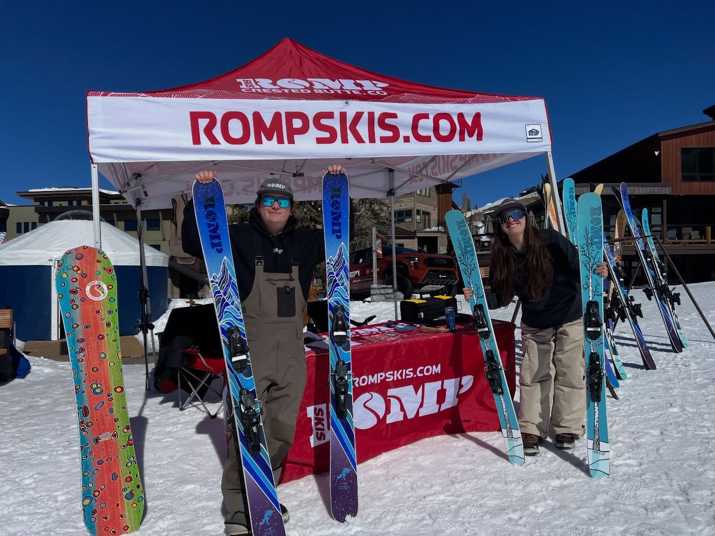 Two people under a "ROMP SKIS" tent on a snowy day, displaying colorful skis. The atmosphere is bright and energetic, with clear blue skies.