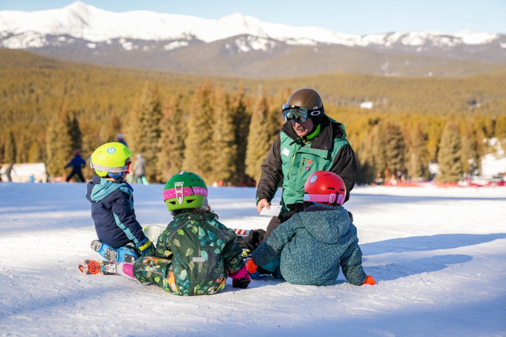 A ski instructor in a green jacket kneels, instructing three children in colorful helmets on a snowy slope, with evergreen trees and mountains in the background.
