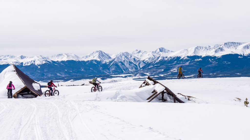 Four cyclists ride fat bikes across a snow-covered landscape with wooden structures. Majestic mountain range in the background under a cloudy sky.