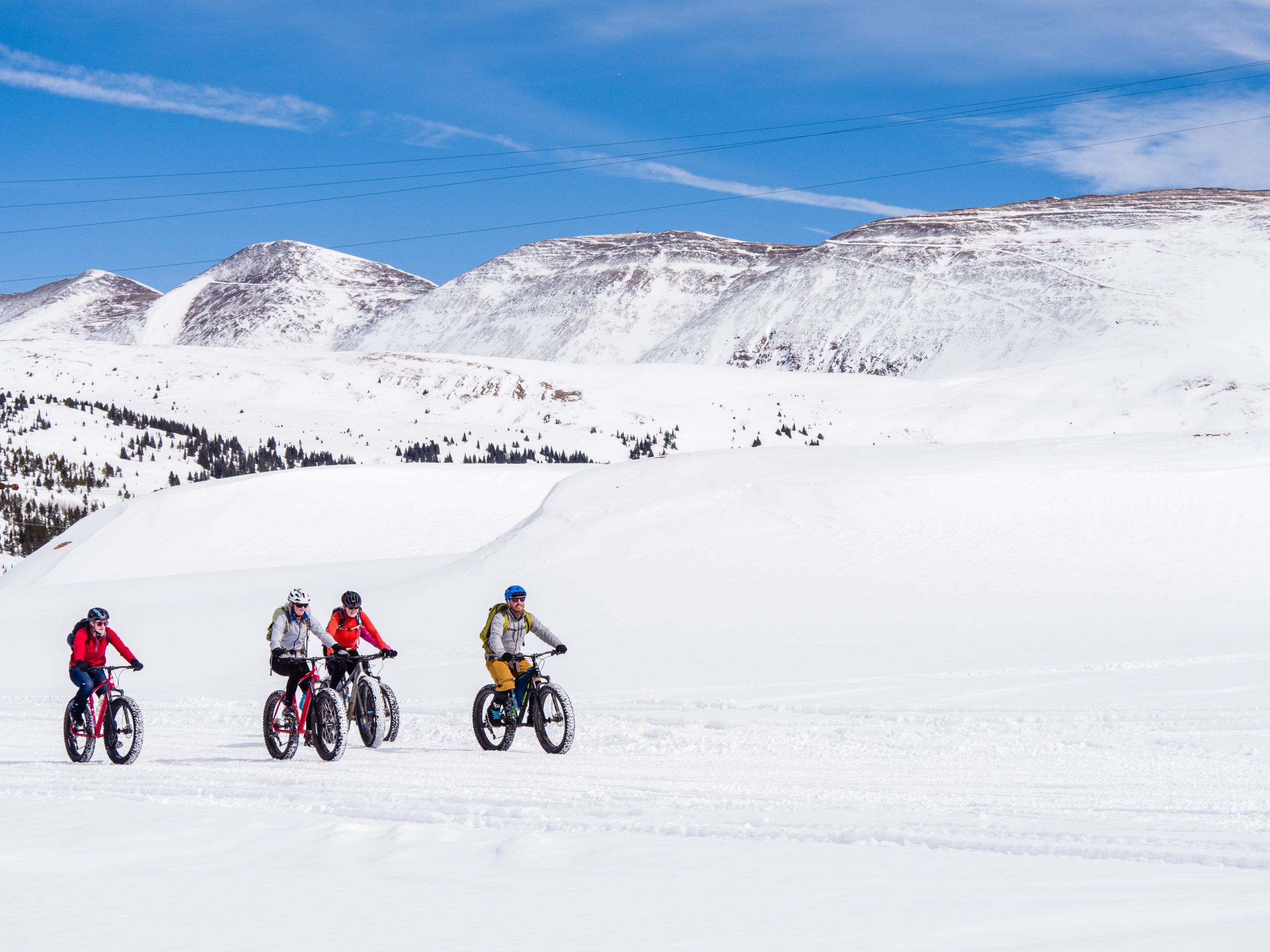 Four people ride fat bikes across a snowy landscape with majestic snow-covered mountains in the background under a clear blue sky.